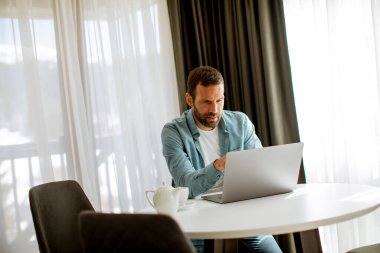 Yakışıklı genç adam dizüstü bilgisayar kullanıyor ve oturma odasında çay içiyor.