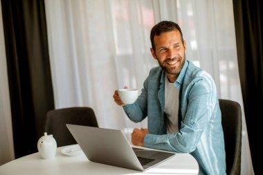 Yakışıklı genç adam dizüstü bilgisayar kullanıyor ve oturma odasında çay içiyor.