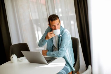 Yakışıklı genç adam dizüstü bilgisayar kullanıyor ve oturma odasında çay içiyor.