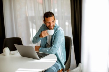 Yakışıklı genç adam dizüstü bilgisayar kullanıyor ve oturma odasında çay içiyor.