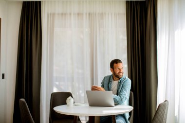 Yakışıklı genç adam dizüstü bilgisayar kullanıyor ve oturma odasında çay içiyor.