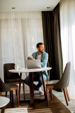Yakışıklı genç adam dizüstü bilgisayar kullanıyor ve oturma odasında çay içiyor.