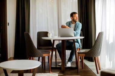 Yakışıklı genç adam dizüstü bilgisayar kullanıyor ve oturma odasında çay içiyor.