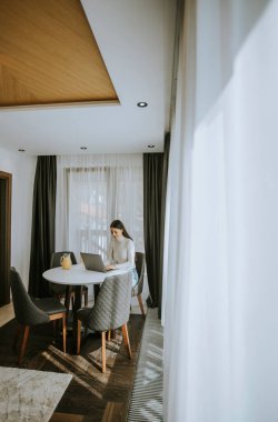 Genç bir kadın dizüstü bilgisayar kullanıyor ve odada sağlıklı portakal suyu içiyor.