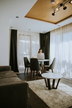 Genç bir kadın dizüstü bilgisayar kullanıyor ve odada sağlıklı portakal suyu içiyor.