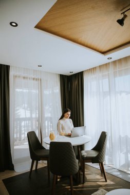 Genç bir kadın dizüstü bilgisayar kullanıyor ve odada sağlıklı portakal suyu içiyor.