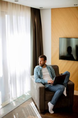 Yakışıklı genç adam oturma odasında dizüstü bilgisayar kullanıyor.