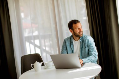 Yakışıklı genç adam dizüstü bilgisayar kullanıyor ve oturma odasında çay içiyor.