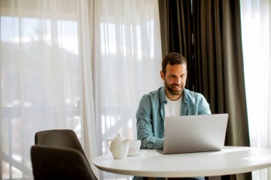 Yakışıklı genç adam dizüstü bilgisayar kullanıyor ve oturma odasında çay içiyor.