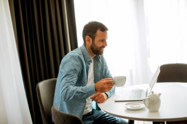 Yakışıklı genç adam dizüstü bilgisayar kullanıyor ve oturma odasında çay içiyor.
