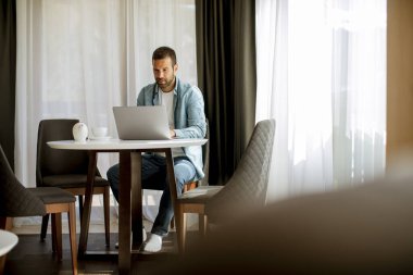 Yakışıklı genç adam dizüstü bilgisayar kullanıyor ve oturma odasında çay içiyor.