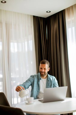 Yakışıklı genç adam dizüstü bilgisayar kullanıyor ve oturma odasında çay içiyor.