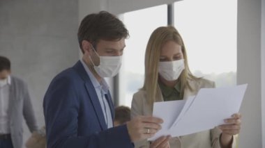 Koruyucu maskeli genç bir adam ve kadın ellerinde kağıtlarla ofisin içinde genç insanlarla tartışıyorlar.