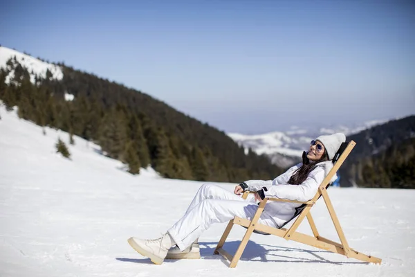 Pretty young woman sitting at deck chair on ski track at the snowy mountain