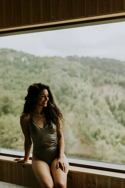 Pretty young woman in wellness spa center sitting by the huge wide window enjoying the green forest view