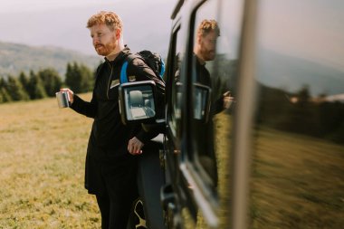 Handsome young man relaxing and drinking coffee by the terrain vehicle hood at countryside
