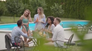 Bir grup mutlu genç içkiyle tezahürat yapıyor ve bahçede havuz başında meyve yiyor.