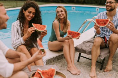 Group of young people sitting by the swimming pool and eating watermelon in the house backyard