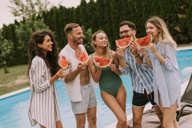 Group of young people standing by the swimming pool and eating watermelon in the house backyard