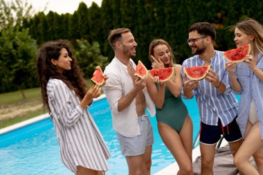 Group of young people standing by the swimming pool and eating watermellon in the house backyard