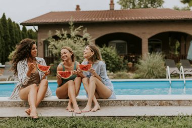 Three cute young women sitting on by the swimming pool and eating watermelon in the house backyard