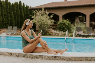 Cute young woman sitting by the swimming pool and listen music from mobile phone with headphones in the house backyard