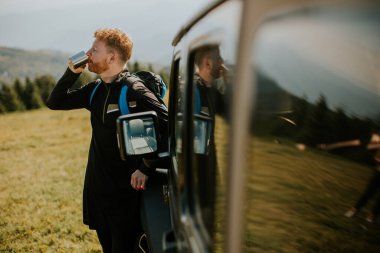Handsome young man relaxing and drinking coffee by the terrain vehicle hood at countryside