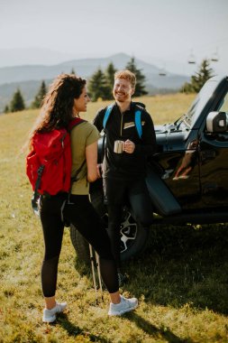 Smiling young couple preparing hiking adventure with backpacks by terrain vehicle