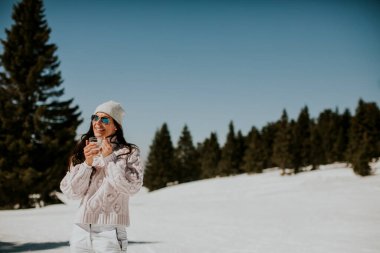 Genç bir kadın güneşli bir kış gününde dağda kayak pistinde limonata içiyor.