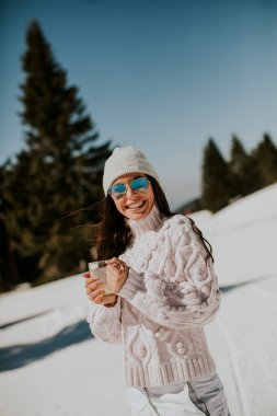 Genç bir kadın güneşli bir kış gününde dağda kayak pistinde limonata içiyor.