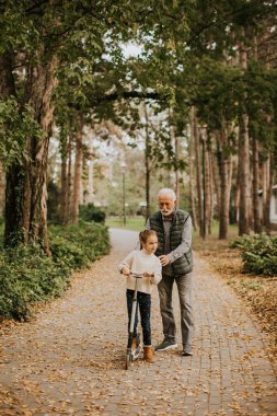 Son sınıf öğrencisi, torununa sonbahar parkında scooter sürmeyi öğretiyor.