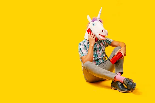 一个戴着独角兽面具的年轻人在电话里说话 用彩色的红色固定电话 坐在黄色的背景上 — 图库照片