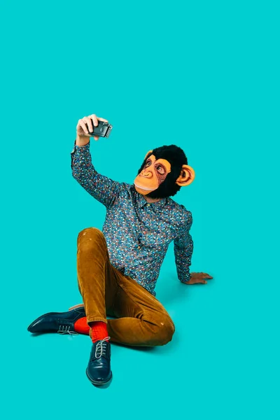 一个戴着猴子面具的年轻人 拿着一个带着复古摄像机的自拍 坐在蓝色的背景上 顶部有一些空白的空间 — 图库照片