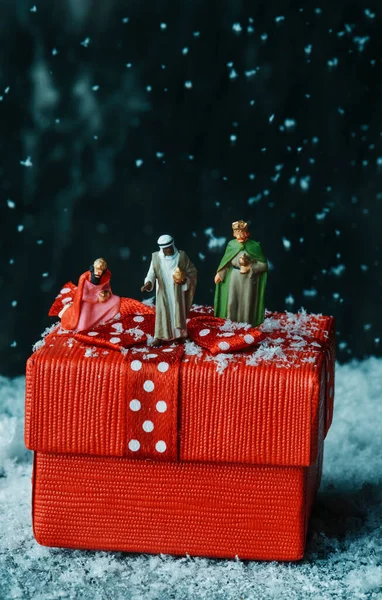 白い点のついたサテンの赤いリボンで結ばれた赤いギフトボックスの上にいる3人の賢者のミニチュアは雪が降る間 — ストック写真