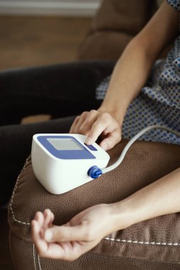 Oturma odasındaki kahverengi koltukta oturan beyaz bir adam kan basıncını elektronik bir sfenometre ile ölçüyor.