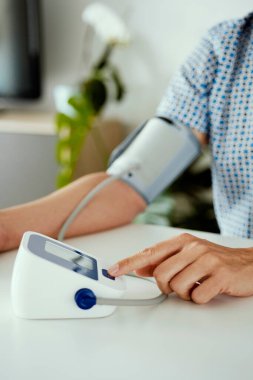 Beyaz bir masada oturan ve kan basıncını elektronik bir sphygmomanometre ile ölçen beyaz bir adamın yakın çekimi.