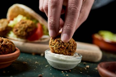Genç bir adam falafeli yoğurt sosuna bandırıyor kırsal yeşil ahşap bir masada oturuyor.