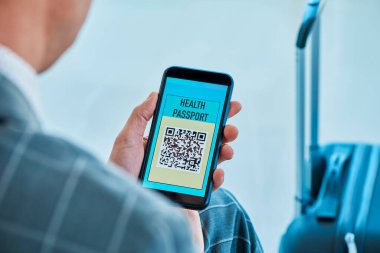 Zarif gri takım elbiseli, genç beyaz bir işadamının yakın plan fotoğrafı. Havaalanının bekleme salonunda oturuyor. Akıllı telefonunun ekranında elektronik sağlık pasaportunun simülasyonunu yapıyor.