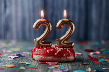 İki altın renkli mum, 22 numarayı oluşturur. Kırmızı şekerli küçük bir pastanın üzerinde, farklı renklerde konfeti serpiştirilmiş gri bir ahşap masa üzerinde.