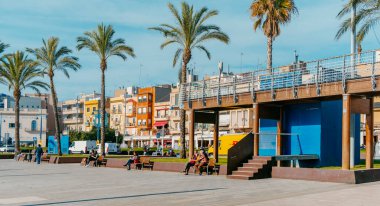 Tarragona, Spain - December 29, 2021: People enjoying the sun on a winter day sitting on the benches of the promenade by the sea at El Serrallo, the traditional fishermen district in Tarragona, Spain