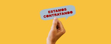 a young man holds a blue paper speech bubble with the text we are hiring written in spanish in it, on a yellow background, in a panoramic format to use as web banner or header