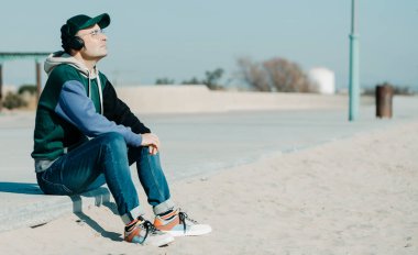 Orta yaşlı bir adam, çok renkli bir kapüşonlu ve kotlu bir adam, güneşli bir kış gününde şehir kumsalının yanında, gezinti alanında oturan bir çift kulaklık kullanarak müzik dinler.