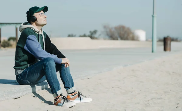 在一个阳光明媚的冬日里 一位穿着五颜六色的帽衫和牛仔裤的中年男子坐在海滨的长廊上 用耳机听音乐 — 图库照片
