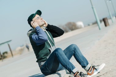 Modern gündelik giysiler giyen olgun bir adam, güneşli bir kış gününde şehir kumsalının yanında, gezinti alanında oturan bir çift kulaklık kullanarak müzik dinliyor.