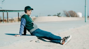 Modern gündelik giysiler giyen orta yaşlı bir adam, güneşli bir kış gününde şehir kumsalının yanında gezinti güvertesinde oturan bir çift kulaklık kullanarak müzik dinliyor.