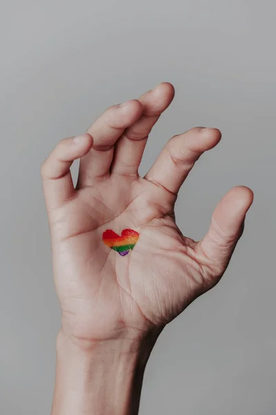 薄い灰色の背景に手のひらにハート型の虹の旗が描かれた男の手のクローズアップ — ストック写真