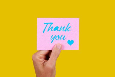 Elinde pembe bir kâğıt parçası olan bir adamın yakın plan fotoğrafı. Üzerinde mavi ile yazılan teşekkür yazısı. Sarı arka plan.