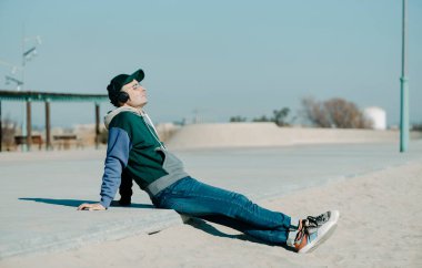 Orta yaşlı bir adam, kot pantolon ve renkli kapüşonlu bir adam, güneşli bir kış gününde şehir plajında yerde oturan bir çift kulaklık kullanarak müzik dinliyor.