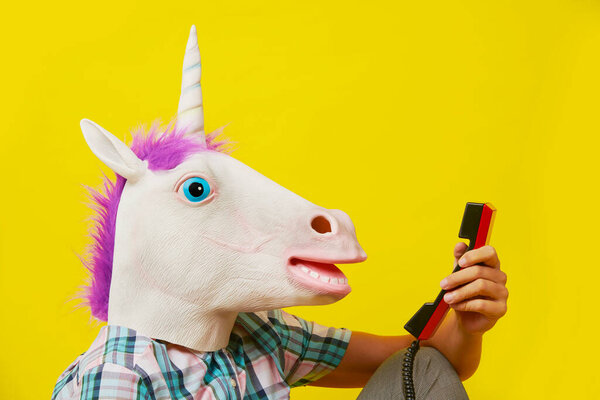 Крупный план головы молодого человека в маске единорога, разговаривающего по красному стационарному телефону, на жёлтом фоне