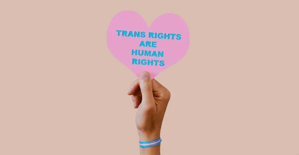 一个年轻的高加索人 戴着印有变性人骄傲旗的腕带 带着一颗粉红的心 上面写着变性人的权利是人权 有着苍白的粉色背景 — 图库照片